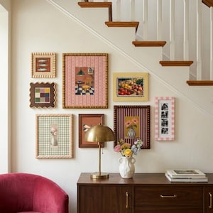 May include: A gallery wall arrangement featuring seven framed art prints with varied patterns and colors, including fish, flowers, and fruit motifs. A brass dome lamp and a white ceramic vase with flowers sit on a wooden sideboard.
