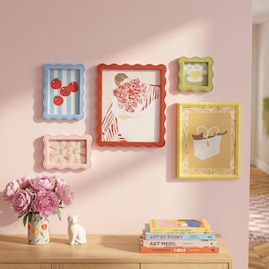 May include: A collection of framed art pieces in various colors and sizes, including a blue frame with tomato art, a red frame with a floral illustration, and a yellow frame with a cat in a bag. A vase of pink flowers and a white cat figurine are on a wooden surface.
