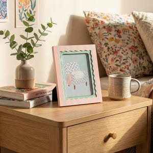 May include: A pink and green striped picture frame with a whimsical illustration of a chair. A small ceramic vase with greenery sits on a stack of books. A ceramic mug rests on a wooden nightstand.