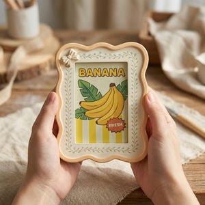 Op de afbeelding: Een cr&egrave;mekleurige, golvende fotolijst met een kleine strik in de linkerbovenhoek. De lijst bevat een grafische print met een tros gele bananen, groene bladeren, gele verticale strepen en de tekst BANANA en FRESH.