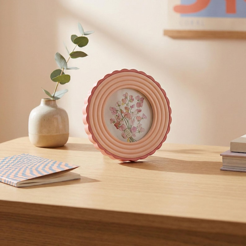 May include: A round, light pink picture frame with a scalloped edge, displaying a floral print. The frame sits on a wooden surface, next to a small vase with greenery and a patterned notebook. The frame's diameter is approximately 6 inches.