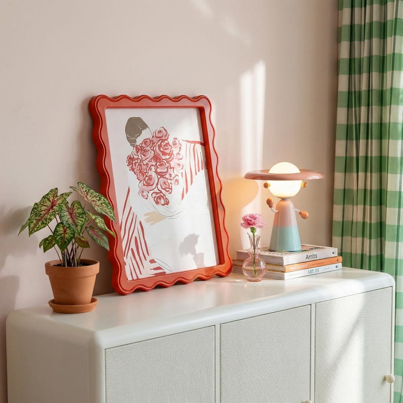 May include: A red framed art print featuring a woman holding roses, displayed on a white cabinet. A decorative lamp with a planet-like design sits nearby, along with a small potted plant and a vase with a pink flower.