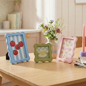 May include: Three decorative picture frames with scalloped edges sit on a wooden table. The frames are blue, green, and pink, each with a unique design. The blue frame has a tomato illustration, the green frame has a teacup, and the pink frame has a floral design.