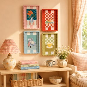 May include: Four rectangular wall frames with scalloped edges in pink, red, light blue, and sage green. The frames contain illustrations of a flower vase, stacked books, an armchair, and a teacup. A table lamp, books, and a ceramic mug are below.