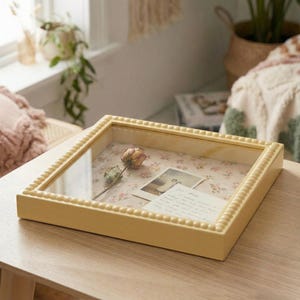 Könnte beinhalten: Ein quadratischer, hellgelber Schattenkasten mit Glasplatte. Im Inneren liegen eine getrocknete Rose, zwei kleine Fotos und eine handgeschriebene Notiz auf einem floral gemusterten Hintergrund. Die Box steht auf einem hellbraunen Holztisch.