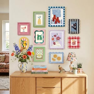 May include: A gallery wall arrangement featuring ten assorted rectangular and oval picture frames in various colors and patterns, including gingham and scalloped edges, surrounding a wooden sideboard with a floral vase and a lamp.