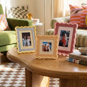 May include: Three decorative photo frames with scalloped edges in yellow, peach, and pink. Each frame holds a photograph with a striped background. The frames are displayed on a wooden coffee table in a living room setting.