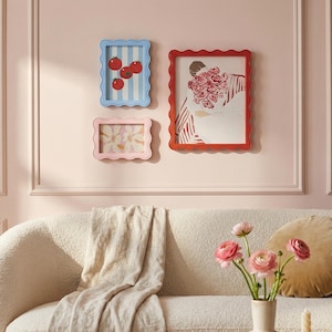 May include: Three framed art pieces with scalloped edges hang on a pink wall. One frame is blue with a tomato illustration, another is pink with a floral print, and the third is red with a person holding flowers.