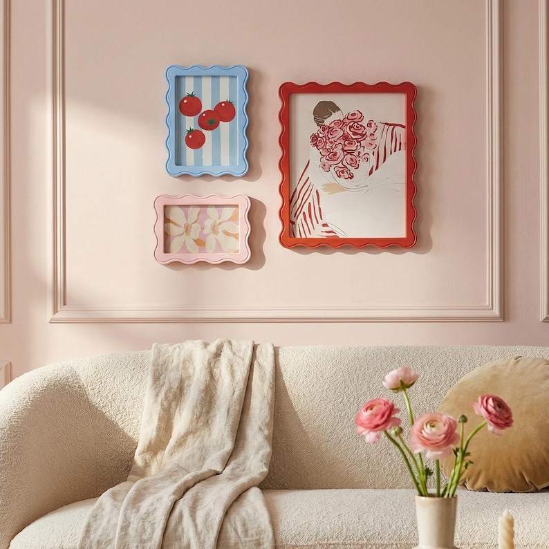 May include: Three framed art pieces with scalloped edges hang on a pink wall. One frame is blue with a tomato illustration, another is pink with a floral print, and the third is red with a person holding flowers.