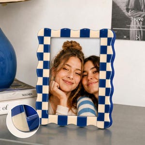 May include: A rectangular picture frame with a wavy, scalloped edge featuring a blue and cream checkered pattern. The frame holds a photograph of two people smiling, and a small circular inset shows a close-up of the frame's corner detail.