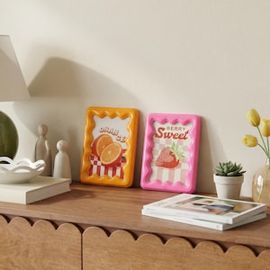 Op de afbeelding: Twee rechthoekige fotolijsten met golvende, geschulpte randen. De linkerlijst is oranje en bevat een print van gesneden sinaasappels met de tekst "ORANGE" op een geblokte achtergrond. De rechterlijst is roze en toont een aardbei met de tekst "BERRY SWEET" op een geblokte achtergrond.