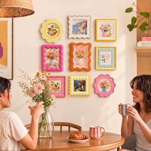 May include: A gallery wall arrangement featuring nine decorative wall frames in various shapes and colors, including scalloped, wavy, and striped designs. The frames contain assorted art prints of floral bouquets, fruit, and line art illustrations.