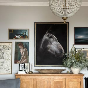 Op de afbeelding: Een verzameling ingelijste kunstwerken en decoraties aan een lichtgrijze muur. De kunst omvat een kaart, een paardenportret, een stilleven en een abstract landschap. Onder de kunst staat een houten kast met een grote witte vaas en groen.