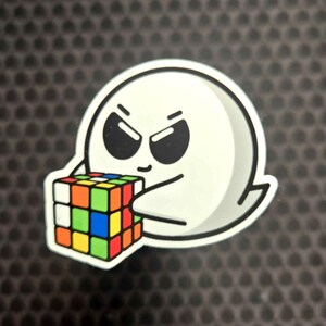 May include: A white ghost-shaped sticker with a black outline, holding a colorful Rubik's Cube. The ghost has black eyes and an angry expression. The cube features red, orange, yellow, green, blue, and white squares.