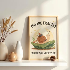 以下が含まれることがあります： 「YOU ARE EXACTLY WHERE YOU NEED TO BE」のテキストが入った額入りのプリントは、葉の上にいるカタツムリの気まぐれなイラストが特徴です。 カタツムリは茶色の殻を持っていて、微笑んでいます。 プリントは明るい木製のフレームに入っており、白い棚に飾られています。