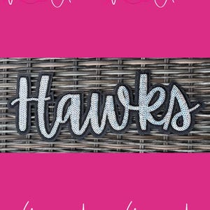 May include: A black patch with the word "Hawks" in silver sequins. The text is outlined in black and sits on a dark background. The background is a woven texture. The image has a pink border with the text "With Andi" repeated.