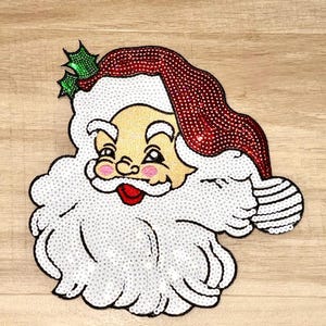 May include: A sequined Santa Claus face patch. The Santa face has a red sequined hat, a white sequined beard, and a smiling face with rosy cheeks. A sprig of green holly is on the hat.