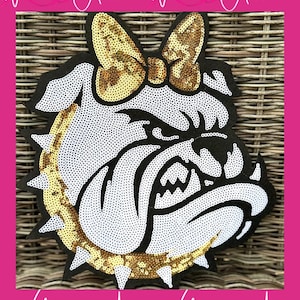 May include: A decorative bulldog head with a bow, made with white and gold sequins on a black background. The bulldog has a fierce expression. The image is framed in pink and includes the text "With Andi" in white script.