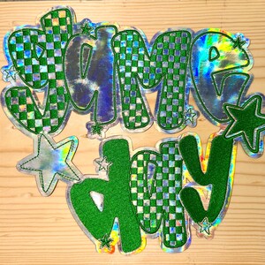 May include: A holographic sticker with the words "GAME DAY" in a bubble font. The letters are outlined in green and filled with a green and white checkered pattern. Several green and silver stars are also included in the design.