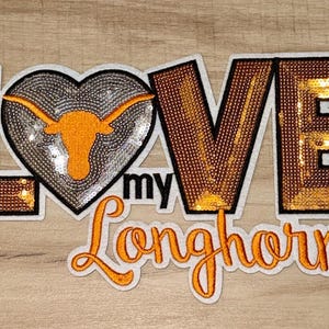 May include: Embroidered patch with the words "LOVE my Longhorns". The letters are embellished with sequins. The heart features a Longhorn silhouette. The patch is outlined in black and white.