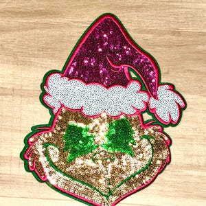 May include: A sequined Christmas ornament in the shape of a green-faced character wearing a Santa hat. The hat is magenta with white trim, and the face is gold with a green bow. The ornament is outlined in green and pink.