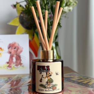 Può includere: Un flacone diffusore a bastoncini in vetro ambrato scuro con un collare color oro e cinque bastoncini beige chiaro. L'etichetta presenta un grafico di una ballerina di hula, un ukulele e un uccello, con il testo Hula Girl e MAKD CANDLES & CO.