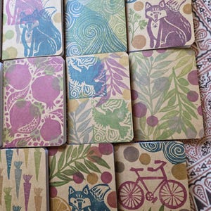 Peut inclure: Plusieurs petits carnets rectangulaires avec des motifs imprimés en bloc sur le thème de la nature. Les motifs comprennent des renards, des vélos, des oiseaux et des motifs botaniques dans des tons de bleu, violet, vert et orange.