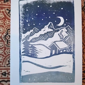 May include: A blue and white linocut print on paper featuring a night landscape with a crescent moon and stars above snow-capped mountains, a small cabin, and evergreen trees. The scene is rendered in a textured, block-printed style.