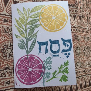 May include: A white card with a colourful print featuring a yellow lemon slice, a pink grapefruit slice, and green foliage. The card also has blue Hebrew text. The background is a patterned fabric.