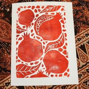 May include: A white card with a red block print design of pomegranates and leaves. The image shows four pomegranates and several leaves in a textured style. The card is set against a patterned fabric, with a white background.