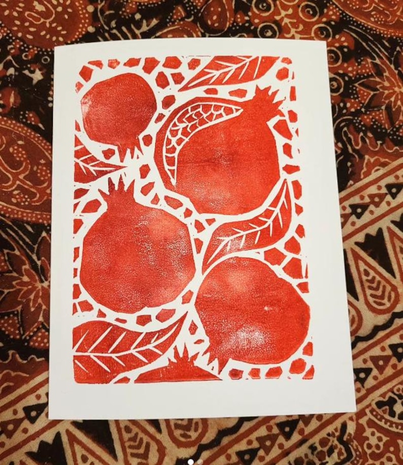 May include: A white card with a red block print design of pomegranates and leaves. The image shows four pomegranates and several leaves in a textured style. The card is set against a patterned fabric, with a white background.