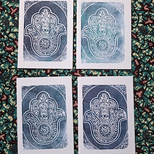 May include: Four white cards featuring a blue and white Hamsa hand design. The Hamsa hand, a symbol of protection, is set within a circular frame. The cards are displayed against a patterned fabric background.