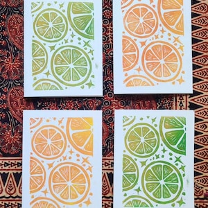 May include: Four rectangular greeting cards featuring block-printed citrus fruit patterns. Two cards display green lime slices with small star accents, while the other two cards feature orange citrus slices with similar star details on white paper.
