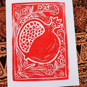 May include: A red and white linocut print of a pomegranate. The fruit is framed by floral and leaf designs. The print is on a white card.