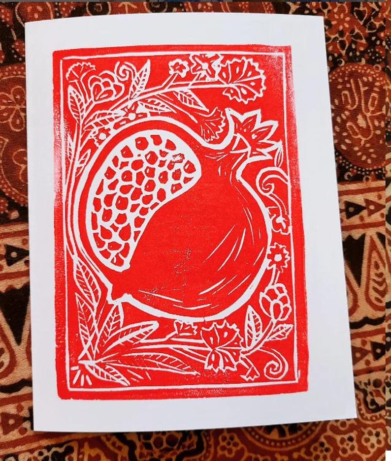 May include: A red and white linocut print of a pomegranate. The fruit is framed by floral and leaf designs. The print is on a white card.