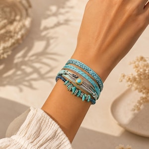 Può includere: Un bracciale a più fili con perline color turchese, un ciondolo in metallo argentato a forma di piuma con una piccola pietra turchese centrale e fasce blu testurizzate effetto pelle. Il design a strati combina diverse texture e dettagli.