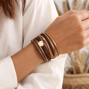 May include: A multi-strand wrap bracelet featuring a combination of brown leather cords, metallic gold-toned chains, and a smooth dark brown textured band. The bracelet is secured with a central circular gold-toned metal charm and a magnetic clasp.