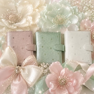 Könnte beinhalten: Drei kleine, rechteckige Planer oder Notizbücher mit Metallic-Finish in zarten Pastelltönen wie Rosa, Mintgrün und Weiß. Jeder Artikel verfügt über ein dezentes Punktemuster, einen vertikalen Riemenverschluss und eine glatte, schimmernde Oberfläche.