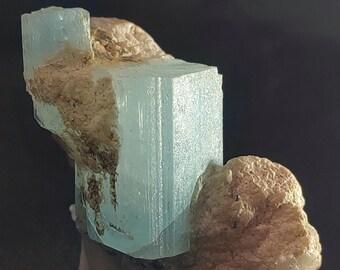May include: A close-up of a raw aquamarine crystal specimen. The light blue crystal is partially embedded in a beige matrix. The crystal has a rectangular shape with visible vertical striations. The background is dark.