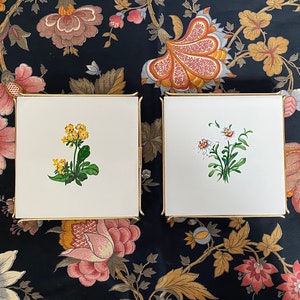 Może przedstawiać: Dwa białe ceramiczne podkładki pod kubki ze złotą obwódką. Jedna podkładka przedstawia żółty kwiat z zielonymi liśćmi. Druga podkładka przedstawia biały kwiat z zielonymi liśćmi.