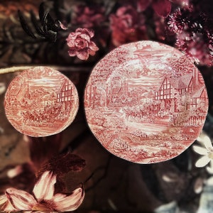 May include: Two vintage ceramic plates with a red and white floral pattern. The larger plate features a countryside scene with a cottage and a horse-drawn carriage. The smaller plate has a similar design with a church and a horse-drawn carriage.