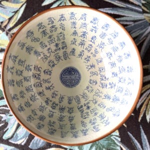 Könnte beinhalten: Eine weiße Keramikschüssel mit blau-weißem Design. Die Schüssel ist mit einem sich wiederholenden Muster chinesischer Schriftzeichen und einem zentralen Symbol verziert.