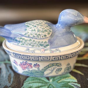 Peut inclure: Un plat en porcelaine bleu et blanc en forme de canard avec un couvercle, décoré de motifs floraux et géométriques. Le plat est orné de fleurs roses, de feuilles vertes et d'une bordure bleu et blanc.