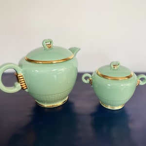 Può includere: Un set vintage di teiera e zuccheriera verde con finiture dorate. La teiera ha un corpo rotondo e un coperchio con un pomo decorativo. La zuccheriera ha una forma simile e un coperchio con un pomo.