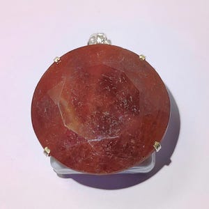 May include: A large, round, reddish-brown gemstone pendant with a faceted surface. The stone is held in place by small silver-coloured metal prongs. A silver-coloured loop is at the top for hanging. The pendant is set against a white background.