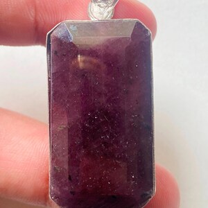 Pode incluir: Um pingente retangular de pedra preciosa facetada em tom roxo profundo, montado em uma estrutura de metal prateado. A pedra apresenta uma superfície plana e polida com inclusões naturais visíveis e um passador prateado decorativo no topo.