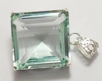 May include: A square-cut, pale green gemstone pendant with a silver bail. The gemstone has multiple facets, creating a sparkling effect. The bail is intricately designed with a decorative pattern. The pendant is set against a white background.