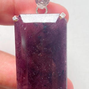 May include: A rectangular, deep purple-red gemstone pendant set in a silver-toned metal prong setting. The stone features a faceted surface with visible natural inclusions and a textured, decorative silver bail at the top for suspension.