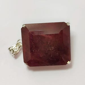 May include: A large, square-cut, deep red gemstone pendant with a silver bail. The gemstone has a faceted surface and is set with small silver prongs at each corner. The bail has a decorative floral design.