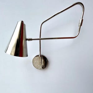 Könnte beinhalten: Eine polierte silberne Wandleuchte mit konischem Schirm und verstellbarem Arm. Die Lampe ist an einer kreisförmigen Basis befestigt und hat ein schlankes, modernes Design. Der Arm der Lampe erstreckt sich und krümmt sich nach oben.